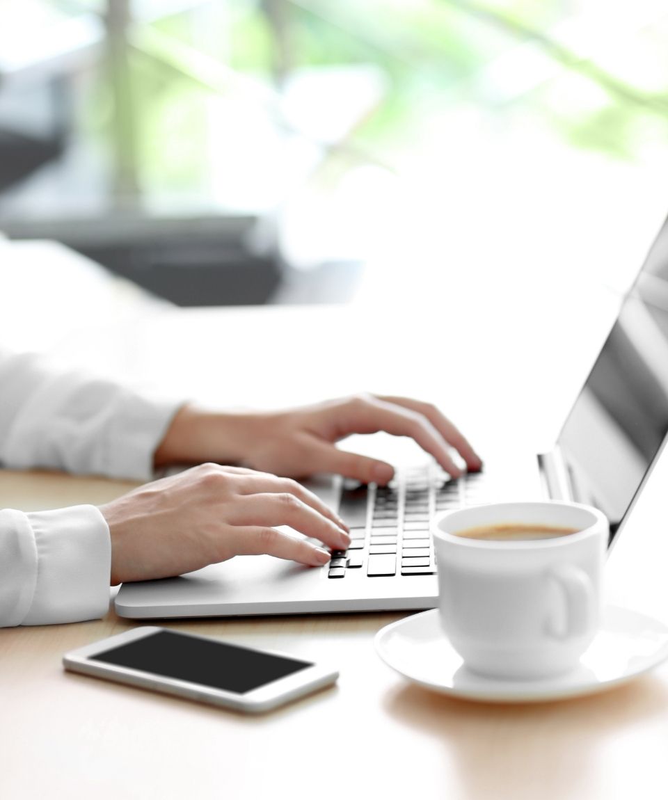 Person's hands shown typing on a laptop. A cell phone and a cup of coffee are next to the laptop in the foreground.